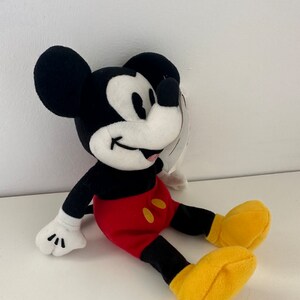 May include: A plush Mickey Mouse toy sitting with a red and yellow outfit. The toy has a black body, white face, and black ears.