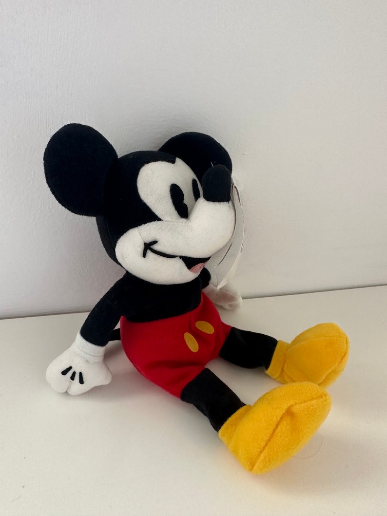 May include: A plush Mickey Mouse toy sitting with a red and yellow outfit. The toy has a black body, white face, and black ears.