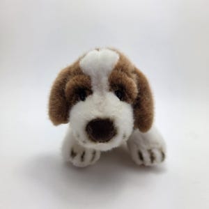 May include: A plush toy dog in a lying position, featuring a brown and white color scheme. The dog has brown ears, a brown nose, and white fur on its face and paws. The toy is designed to resemble a puppy.