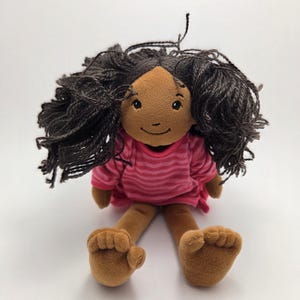 Puede incluir: Una muñeca de peluche con piel morena, cabello rizado oscuro y un vestido rosa a rayas. La muñeca tiene pies y brazos marrones y una cara sonriente. La muñeca está sentada sobre una superficie blanca.
