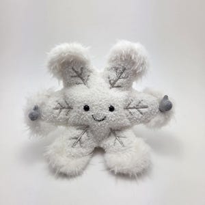 Puede incluir: Un juguete de peluche blanco con forma de copo de nieve con una cara sonriente y detalles bordados en gris. El copo de nieve tiene seis brazos esponjosos y pequeños pulgares grises hacia arriba. El juguete está diseñado para parecerse a un copo de nieve amigable.
