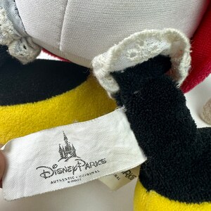 K&ouml;nnte beinhalten: Ein gelbes, schwarzes und rotes Pl&uuml;schtier mit einer wei&szlig;en Spitzenborte. Das Spielzeug hat ein Etikett mit der Aufschrift "Disney Parks Authentic Original".