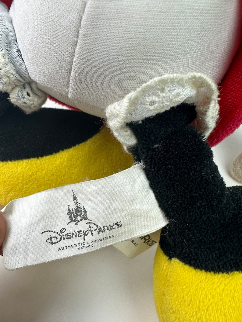 K&ouml;nnte beinhalten: Ein gelbes, schwarzes und rotes Pl&uuml;schtier mit einer wei&szlig;en Spitzenborte. Das Spielzeug hat ein Etikett mit der Aufschrift "Disney Parks Authentic Original".