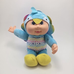 May include: A blue plush Cabbage Patch Kid doll wearing a blue and yellow bird costume with a rainbow tail. The doll has a smiling face with blue eyes and a yellow beak.