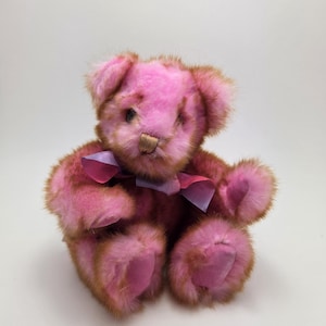 May include: A pink teddy bear with brown accents. The plush toy has a pink bow around its neck and is sitting upright. The bear's fur is a soft, textured material, and it has a stitched nose and small, dark eyes.