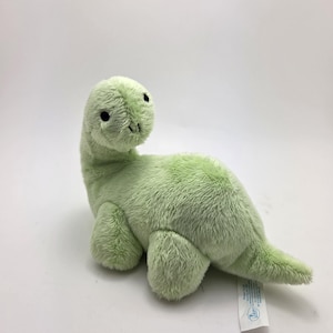 Könnte beinhalten: Ein hellgrünes Plüsch-Dinosaurier-Spielzeug mit langem Hals, kleinen schwarzen Augen und kurzen Beinen. Das Stofftier besteht aus einem flauschigen Material und hat ein kleines Etikett an seinem Schwanz. Der Dinosaurier liegt auf einer weißen Oberfläche.
