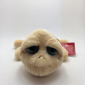 May include: A plush turtle toy with a tan body, large green eyes, and a sad expression. The toy has a tag that reads "RUSS Make someone happy."