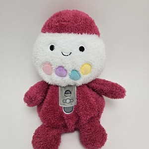 May include: A plush toy shaped like a gumball machine with a red body, white head, and a gray dispenser. The head has a smiling face and colorful gumballs.