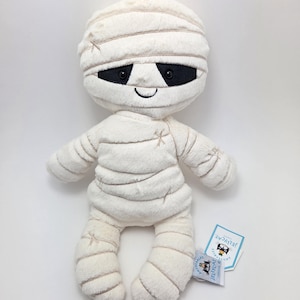 Op de afbeelding: Wit pluche mummiespeelgoed met zwarte ogen en een glimlachende mond. Het speelgoed draagt een witte bandageachtige outfit met bruine stiksels. Het speelgoed heeft een label met de tekst "Jellycat" erop.