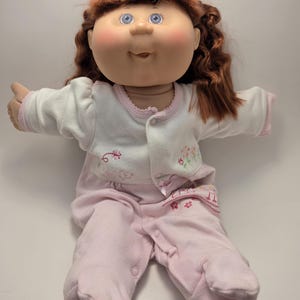 May include: A Cabbage Patch Kid doll with auburn hair and blue eyes, wearing a white jacket with pink trim and a pink onesie. The doll has rosy cheeks and is seated with arms outstretched.