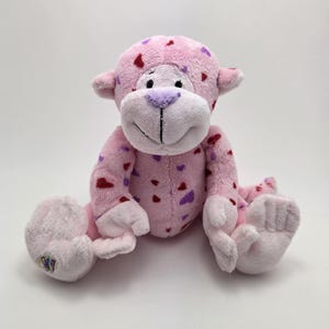 May include: A pink plush monkey toy with red and purple heart patterns. The soft toy has a white face, purple nose, and is sitting with its legs spread. A children's toy.