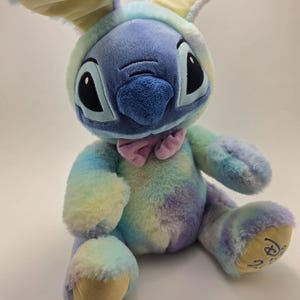 May include: A plush toy of the character Stitch, dressed in a pastel rainbow bunny costume. The toy has large black eyes, a blue nose, and a pink bow tie. The bunny ears are yellow and blue.