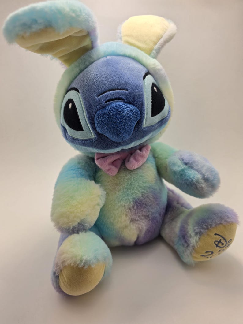 May include: A plush toy of the character Stitch, dressed in a pastel rainbow bunny costume. The toy has large black eyes, a blue nose, and a pink bow tie. The bunny ears are yellow and blue.