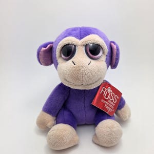 May include: Purple plush monkey with large, glittery eyes. The monkey is sitting with its legs crossed and has a red tag that says "Russ Make someone happy".