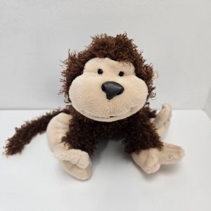 May include: A brown and beige plush monkey toy with a furry texture. The monkey is sitting with its legs crossed and has a friendly expression.