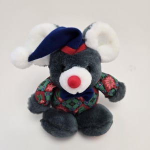 May include: A gray plush mouse wearing a blue and red plaid shirt with a red nose and a blue sleeping cap. The mouse is sitting on a white background.