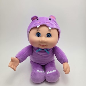 May include: A Cabbage Patch Kids doll wearing a purple hippo costume. The doll has blue eyes, a small smile, and a light skin tone. The hippo costume includes a hood with ears and teeth, and the doll is sitting with its arms outstretched.