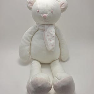 Könnte beinhalten: Ein weißer Plüsch-Teddybär mit einer strukturierten Stoffoberfläche. Der Bär hat hellrosa gestickte Augen, eine rosa Nase und einen passenden rosa gestickten Mund. Er trägt einen weißen Schal mit kleinen rosa Punkten um den Hals.
