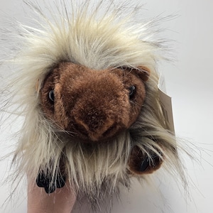 May include: A plush porcupine toy featuring a soft brown face with dark eyes and a prominent nose. The body is covered in long, shaggy, cream-colored faux fur that mimics quills, with small black paws visible at the front and a white tag attached.