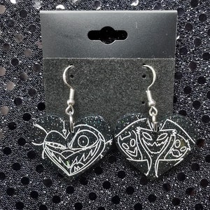 May include: A pair of black heart-shaped earrings with white glitter and a white line drawing of a character from the movie Nightmare Before Christmas. The earrings are hanging from silver hooks.