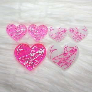 May include: Five heart-shaped resin earrings with a white cartoon character design. Two of the earrings are pink with glitter, and three are white.