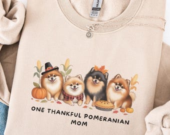 May include: Sand-colored sweatshirt featuring a graphic of four Pomeranian dogs with fall-themed accessories. The text "ONE THANKFUL POMERANIAN MOM" is printed below the dogs. The sweatshirt is paired with blue jeans.