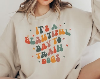 May include: A beige sweatshirt with a colorful retro-style graphic that says "It's a beautiful day to train dogs".