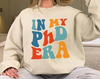 May include: A cream colored sweatshirt with the text "IN MY PHD ERA" in colorful retro font.