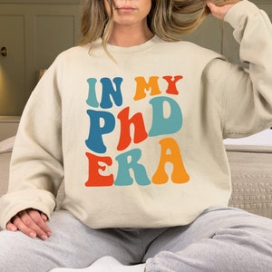 May include: A cream colored sweatshirt with the text "IN MY PHD ERA" in colorful retro font.