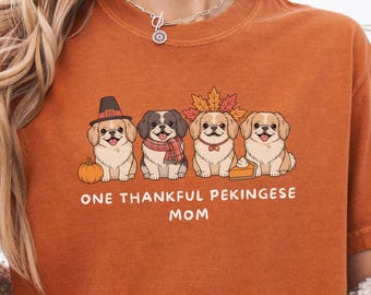 May include: Burnt orange t-shirt featuring a graphic of four Pekingese dogs with fall-themed accessories. The dogs are wearing a pilgrim hat, scarf, and fall leaves. The text "ONE THANKFUL PEKINGESE MOM" is printed below the dogs.