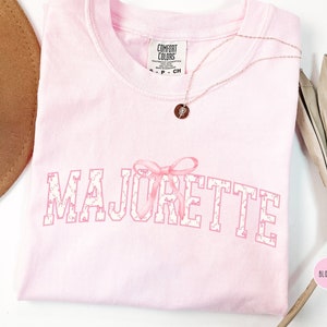 May include: Pink t-shirt with the word "MAJORETTE" in white letters with a pink bow and a white polka dot pattern. The shirt is on a white surface.
