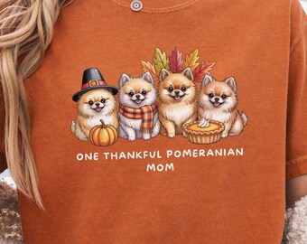 May include: Burnt orange t-shirt featuring a graphic of four Pomeranian dogs with fall-themed accessories. The dogs are surrounded by autumn leaves and a pumpkin pie. Text reads "ONE THANKFUL POMERANIAN MOM".