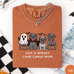 May include: A rust-coloured t-shirt with a Halloween graphic of four Cane Corso dogs in costumes: a ghost, vampire, mummy, and holding jack-o'-lanterns. The text reads "JUST A SPOOKY CANE CORSO MOM". A silver chain necklace is draped across the shirt.