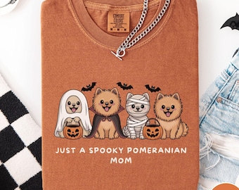 May include: A burnt orange t-shirt featuring a Halloween-themed graphic. The design includes four Pomeranian dogs dressed as a ghost, vampire, mummy, and a regular dog, with bats and pumpkins. The text reads "JUST A SPOOKY POMERANIAN MOM".