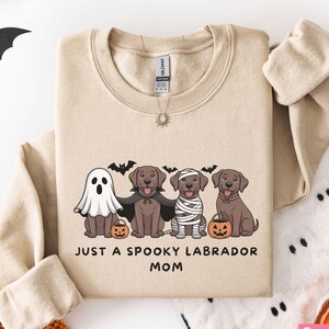 May include: Sand-colored sweatshirt with a Halloween-themed graphic. The design features four Labrador retrievers dressed as a ghost, vampire, mummy, and a dog with a pumpkin. The text reads "JUST A SPOOKY LABRADOR MOM".