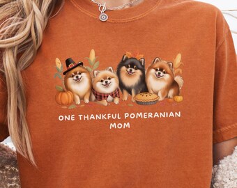 May include: Burnt orange t-shirt featuring a graphic of four Pomeranian dogs with fall-themed accessories. The text "ONE THANKFUL POMERANIAN MOM" is printed below the dogs. The shirt has a crew neck and short sleeves.