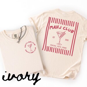 Puede incluir: Dos camisetas color marfil con gr&aacute;ficos rojos. Una tiene "EMILY'S MAHJ CLUB" y una copa de c&oacute;ctel. La otra tiene "MAHJ CLUB", una copa de c&oacute;ctel, "EST 2026" y "CRACKS, DOTS, BAMS". Una cadena de plata est&aacute; en la camiseta delantera. La palabra "ivory" est&aacute; escrita en negro.