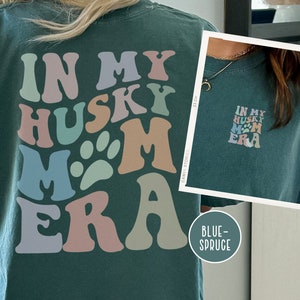May include: A dark green t-shirt with a retro-style graphic that says "In My Husky Mom Era" in colorful letters and a paw print. The shirt is shown on a model.