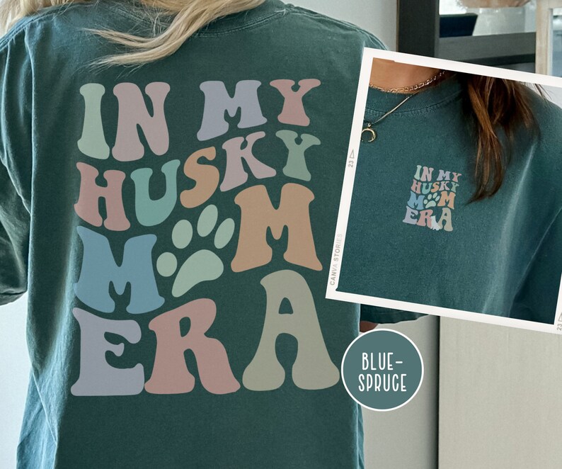 May include: A dark green t-shirt with a retro-style graphic that says "In My Husky Mom Era" in colorful letters and a paw print. The shirt is shown on a model.