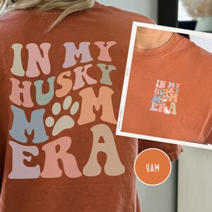 May include: A rust-colored t-shirt with the text "In My Husky Mom Era" in a retro font with paw prints. The text is in shades of pink, blue, green, and beige.