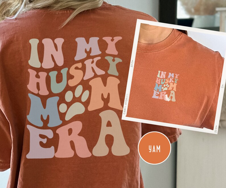 May include: A rust-colored t-shirt with the text "In My Husky Mom Era" in a retro font with paw prints. The text is in shades of pink, blue, green, and beige.