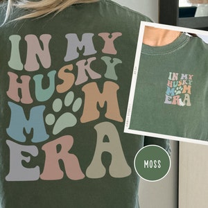 May include: A green t-shirt with the text "In My Husky Mom Era" in colorful, retro-style lettering. The text includes a paw print graphic.