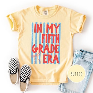 May include: A pale yellow short-sleeve t-shirt featuring a graphic with vertical blue stripes and the red text "IN MY FIFTH GRADE ERA" with a small red bow illustration. The shirt is displayed alongside a pair of black and white chequered slip-on shoes.