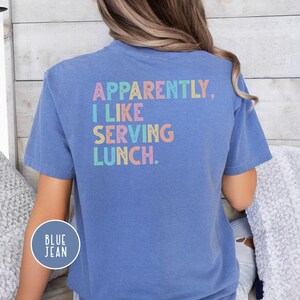 May include: A blue t-shirt with the text "APPARENTLY, I LIKE SERVING LUNCH" in pastel colors. The shirt has short sleeves and a round neckline. A circular "BLUE JEAN" label is on the lower left side of the shirt.