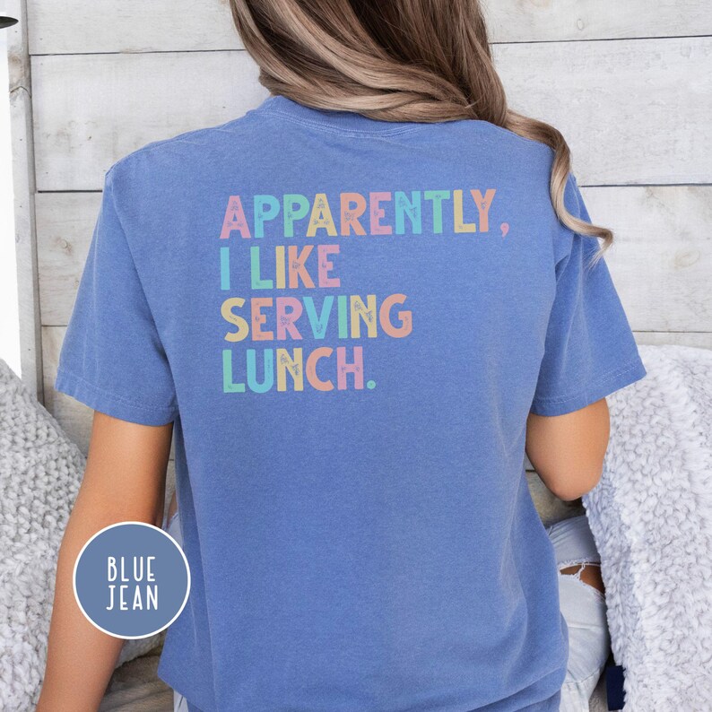 May include: A blue t-shirt with the text "APPARENTLY, I LIKE SERVING LUNCH" in pastel colors. The shirt has short sleeves and a round neckline. A circular "BLUE JEAN" label is on the lower left side of the shirt.
