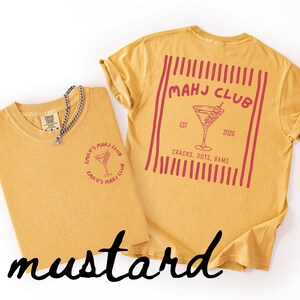 Puede incluir: Camisetas de color mostaza con un gr&aacute;fico de c&oacute;ctel. Una camiseta tiene un logotipo peque&ntilde;o, la otra tiene un dise&ntilde;o m&aacute;s grande con el texto "MAHJ CLUB" y "CRACKS, DOTS, BAMS". Una cadena de plata est&aacute; en una camiseta.