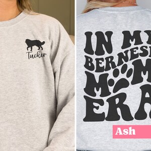 May include: A grey sweatshirt with a black silhouette of a Bernese Mountain Dog and the name "Tucker" on the front. The back of the sweatshirt has a black graphic that says "In My Bernese Mom Era" with a paw print in the middle. The name "Ash" is printed in pink at the bottom of the sweatshirt.