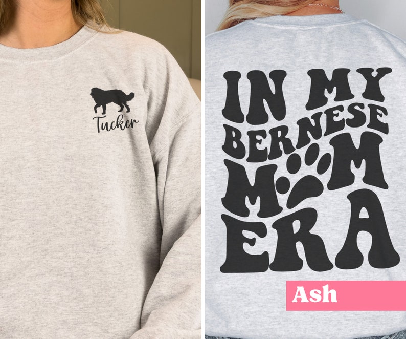 May include: A grey sweatshirt with a black silhouette of a Bernese Mountain Dog and the name "Tucker" on the front. The back of the sweatshirt has a black graphic that says "In My Bernese Mom Era" with a paw print in the middle. The name "Ash" is printed in pink at the bottom of the sweatshirt.
