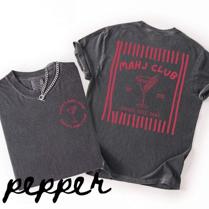 Puede incluir: Dos camisetas grises oscuras con gr&aacute;ficos rojos. Una camiseta tiene un dise&ntilde;o de copa de c&oacute;ctel con el texto "EMILY'S MAHJ CLUB". La otra camiseta tiene un dise&ntilde;o similar en la espalda con el texto "MAHJ CLUB" y "2026". Una cadena de plata est&aacute; en una camiseta.