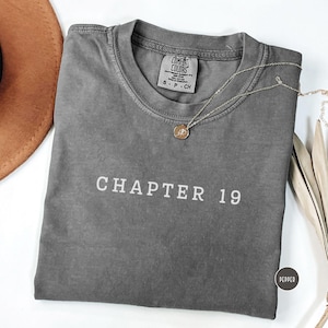 Puede incluir: Una camiseta gris Comfort Colors con el texto "CHAPTER 19" en blanco. Un collar plateado con un colgante circular descansa sobre la camiseta. La camiseta está doblada y tiene una pequeña etiqueta "PEPPER".